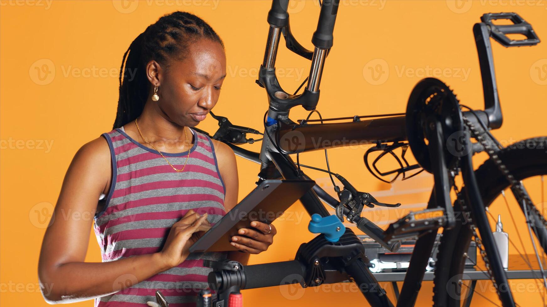 African american woman following online tutorials on tablet, learning to repair her damaged bike. Cycling hobbyist following instructions on how to do upkeeping for bicycle parts, studio background photo