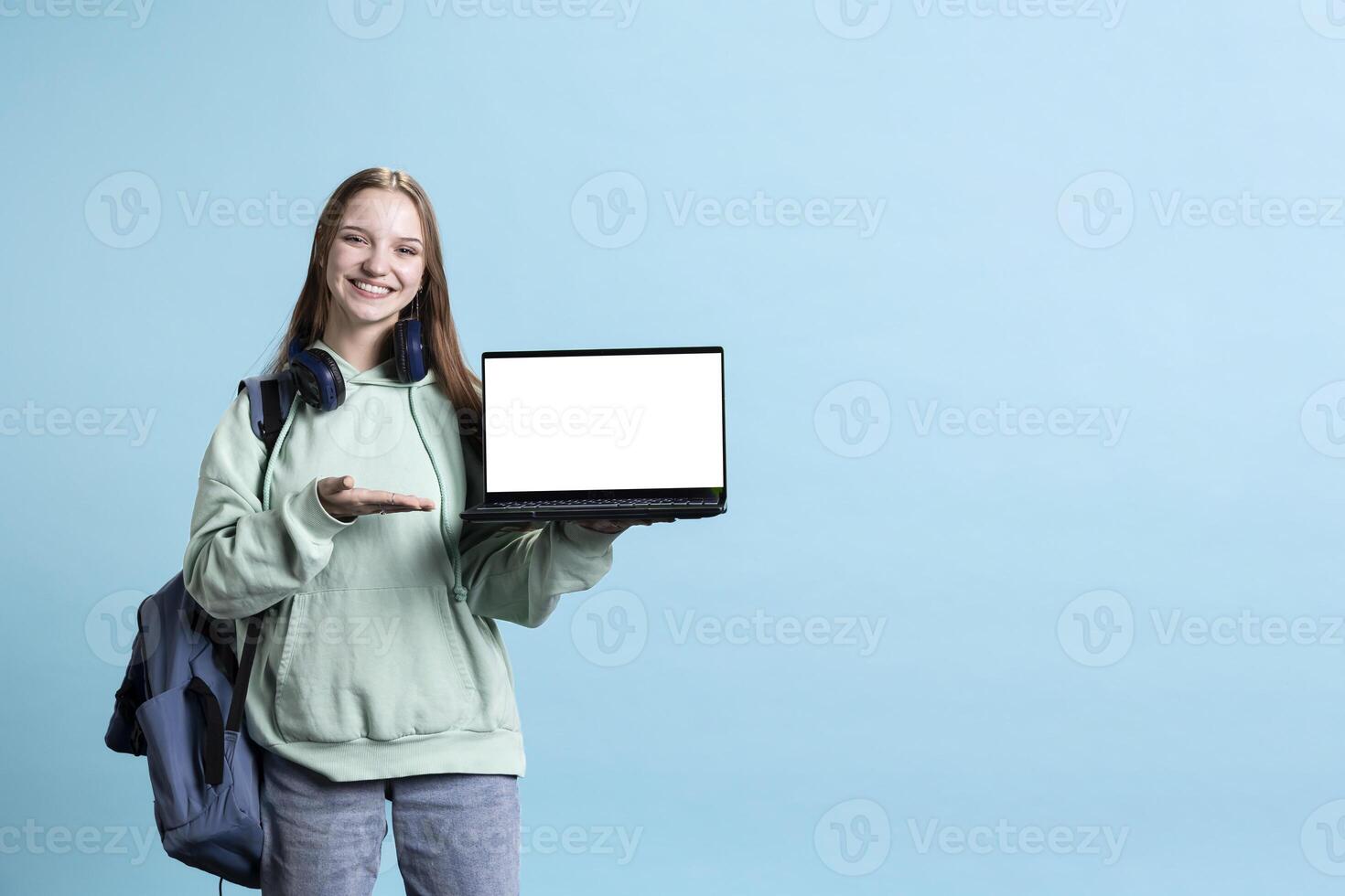 Upbeat girl pointing towards laptop with isolated screen display, doing advertisement, studio background. Radiant teenager grinning, creating ad promotion with mockup notebook device photo