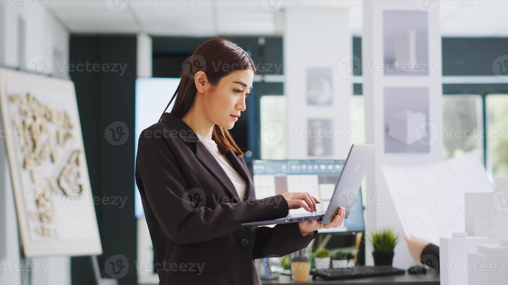 Developer makes sketches on laptop for construction program while inspecting 3D building model on design table in coworking space. Contractor developing renovation scheme and generating blueprint. photo