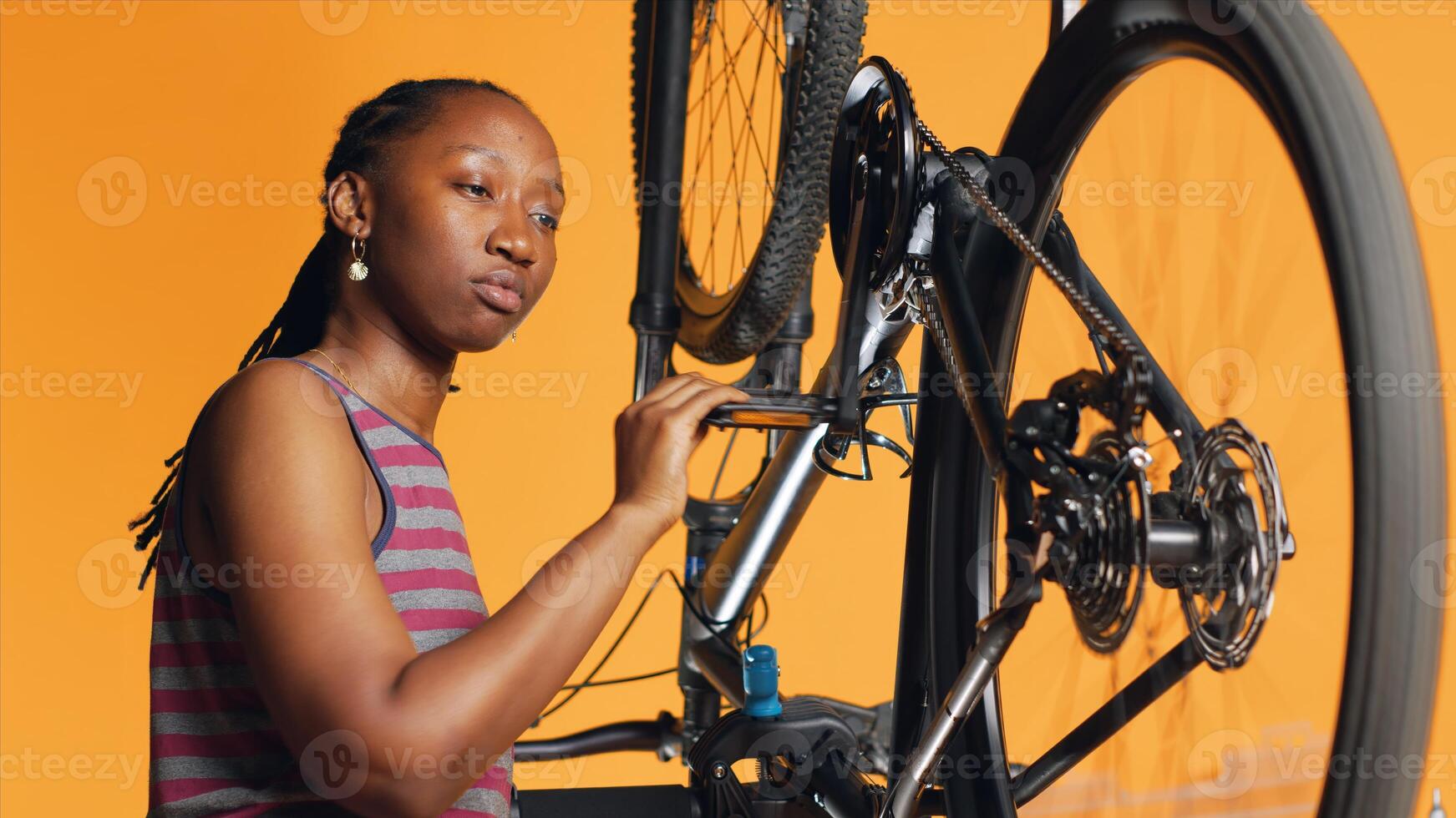 BIPOC engineer checking bicycle performance by spinning pedals. Skilled specialist ensuring proper function of tires on bike wheels in orange studio background during maintenance, camera A photo