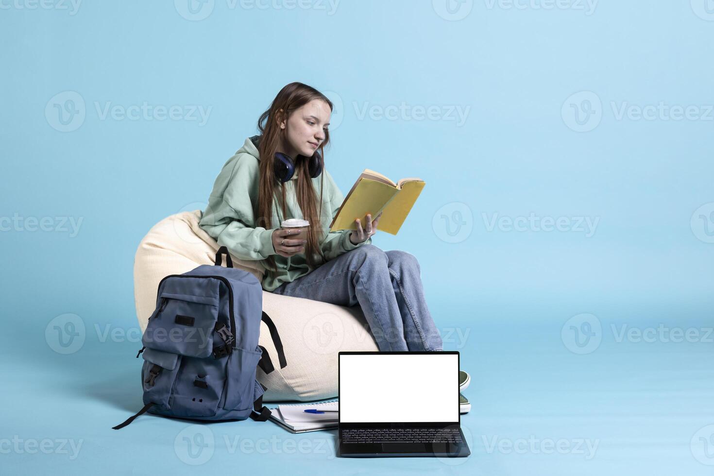 Mockup laptop next to schoolgirl holding coffee cup and reading textbook, preparing for university exams, studio background. Teenager consuming beverage, enjoying book next to isolated screen notebook photo