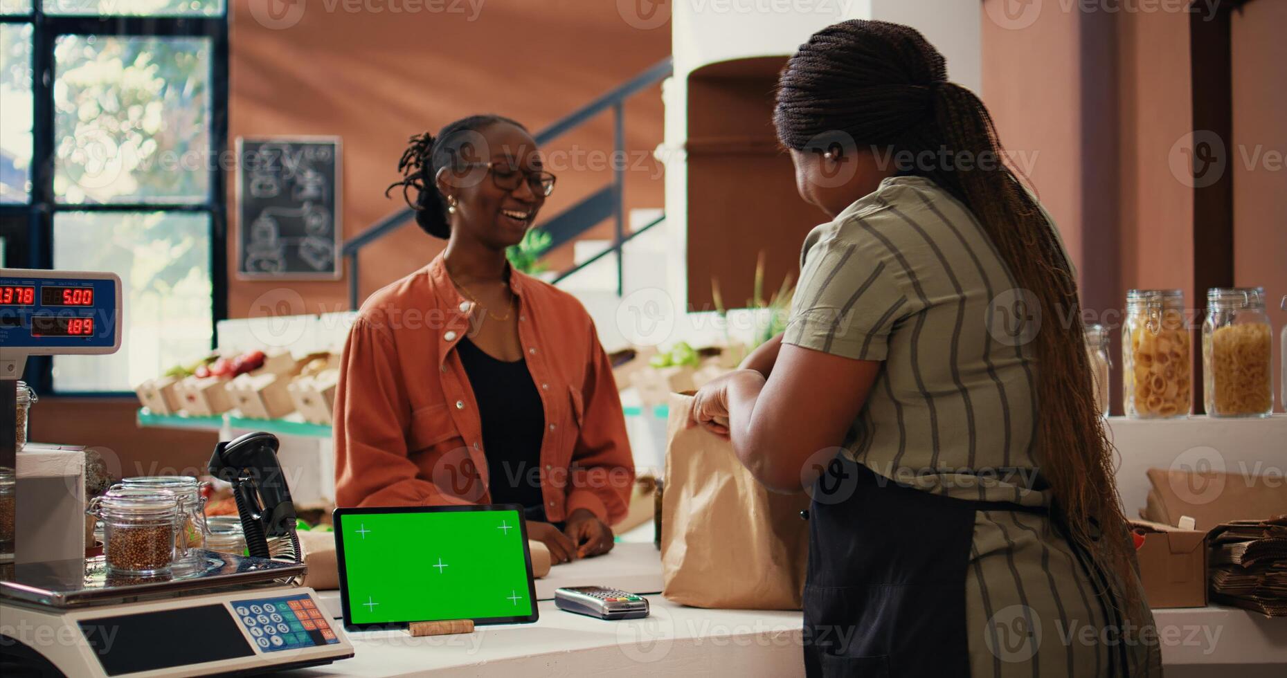 Customer approaching counter with greenscreen on device, buying eco friendly goods from local owner. African american vendor using isolated copyspace display on tablet, talks to client. Tripod shot. photo