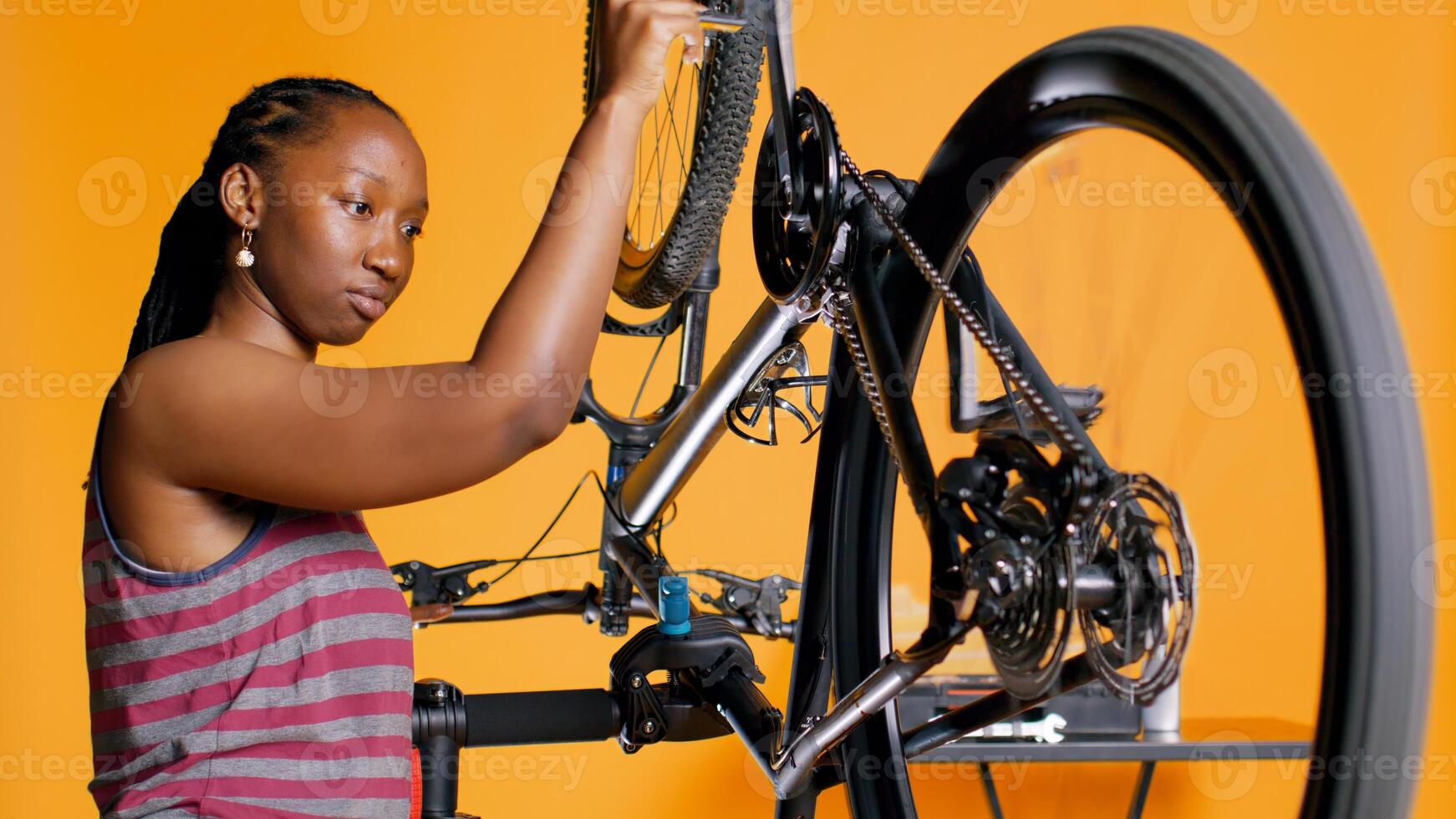 African american woman checking bicycle performance by spinning pedals. Professional ensuring proper function of tires on bike wheels in orange studio background, camera B photo