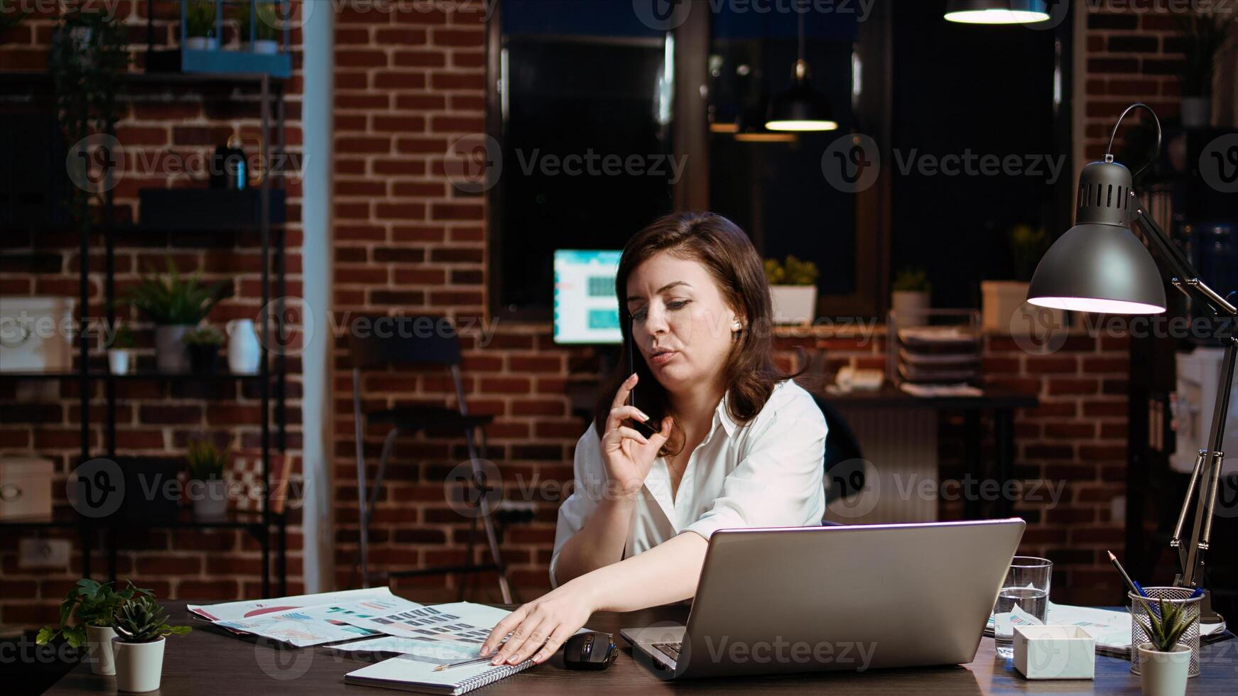 Woman answers phone call from husband while working in office at computer desk. Employee using smartphone to chat with friend during telephone call while at work in front of laptop at night, camera A photo