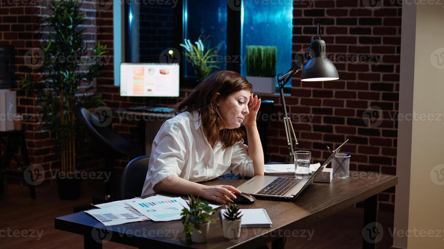 Businesswoman working overtime for company project, doing tasks in office overnight, struggling to remain awake. Tired employee yawning while looking over data on laptop screen late at night, camera A photo