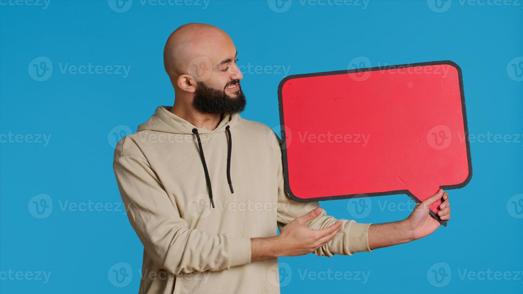 Arab person holding red speech bubble icon in studio, creating web advertisement for new product commercial. Middle eastern man shows isolated cardboard sign for ad. Camera 1. photo