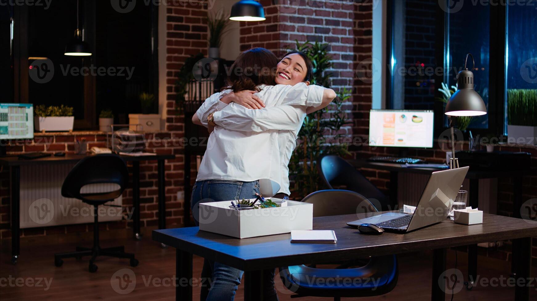 Woman packing office desk belongings, excited to go on holiday, hugging asian coworker. Joyful employee putting objects on workspace table in box, taking days off to go on vacation, camera A photo