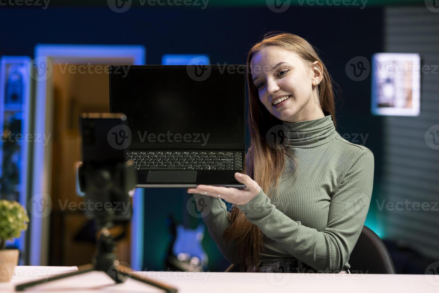 Happy gen Z online show host filming content about best laptop releases of the year with smartphone on tripod. Upbeat young influencer recording newly launched notebook review using mobile phone photo