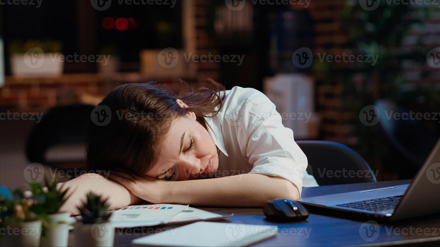 Sleepy employee asleep on computer desk late at night in brick wall office. Panning shot of tired businesswoman sleeping with head on table while working overnight on laptop, camera B photo
