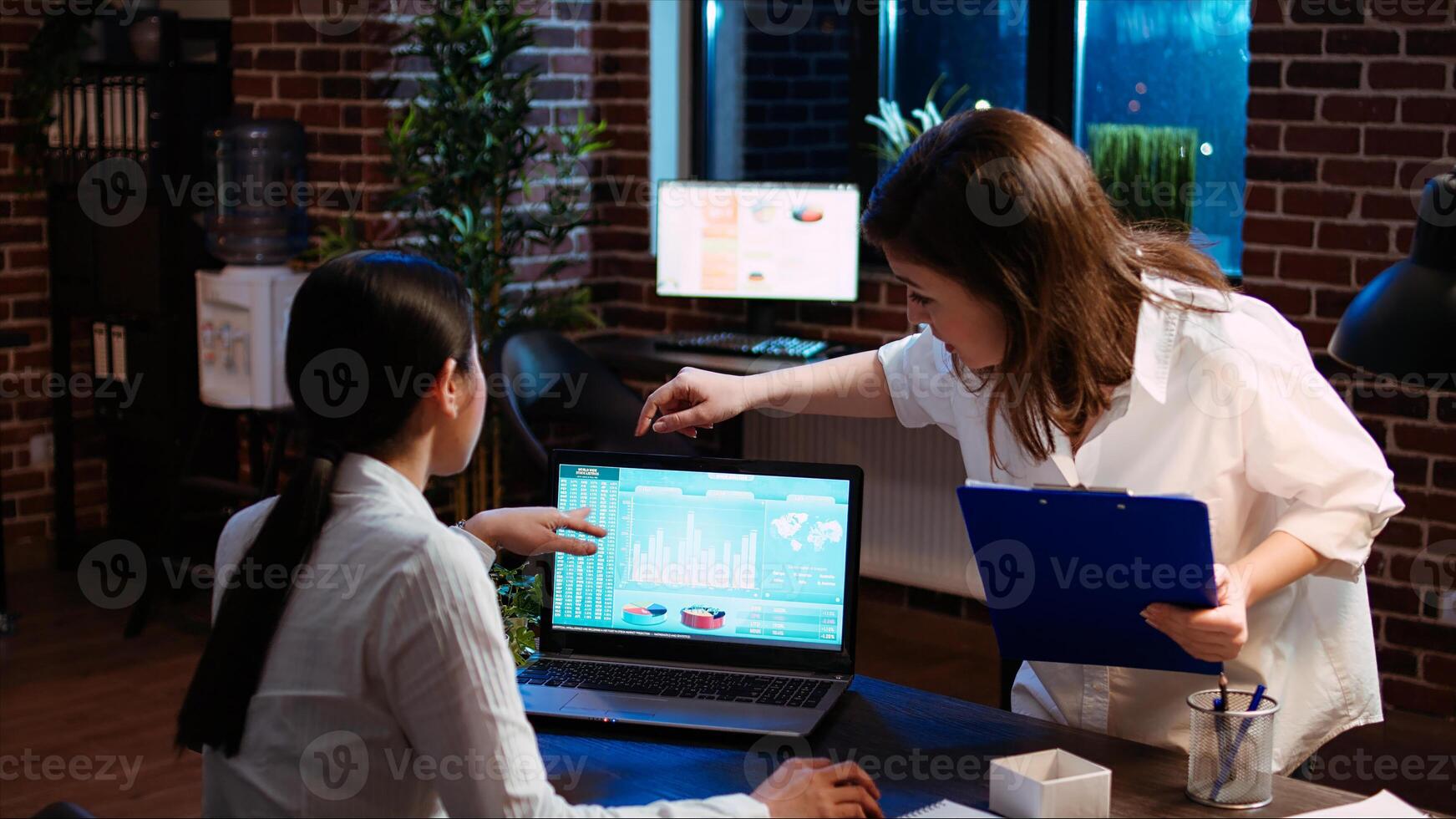 Company accountant and coworker analyzing financial data graphs on laptop screen. Employee and colleague comparing fourth quarter revenue figures in brick wall office, camera A photo