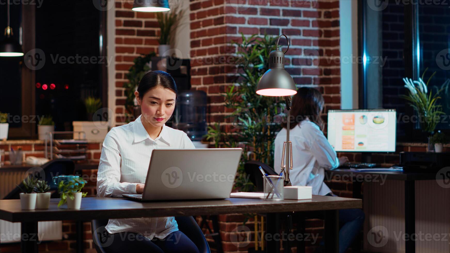 Smiling asian worker researching key data for company project, doing tasks in office overnight. Cheerful staff member looking over accounting figures on laptop screen late at night, typing on keyboard photo