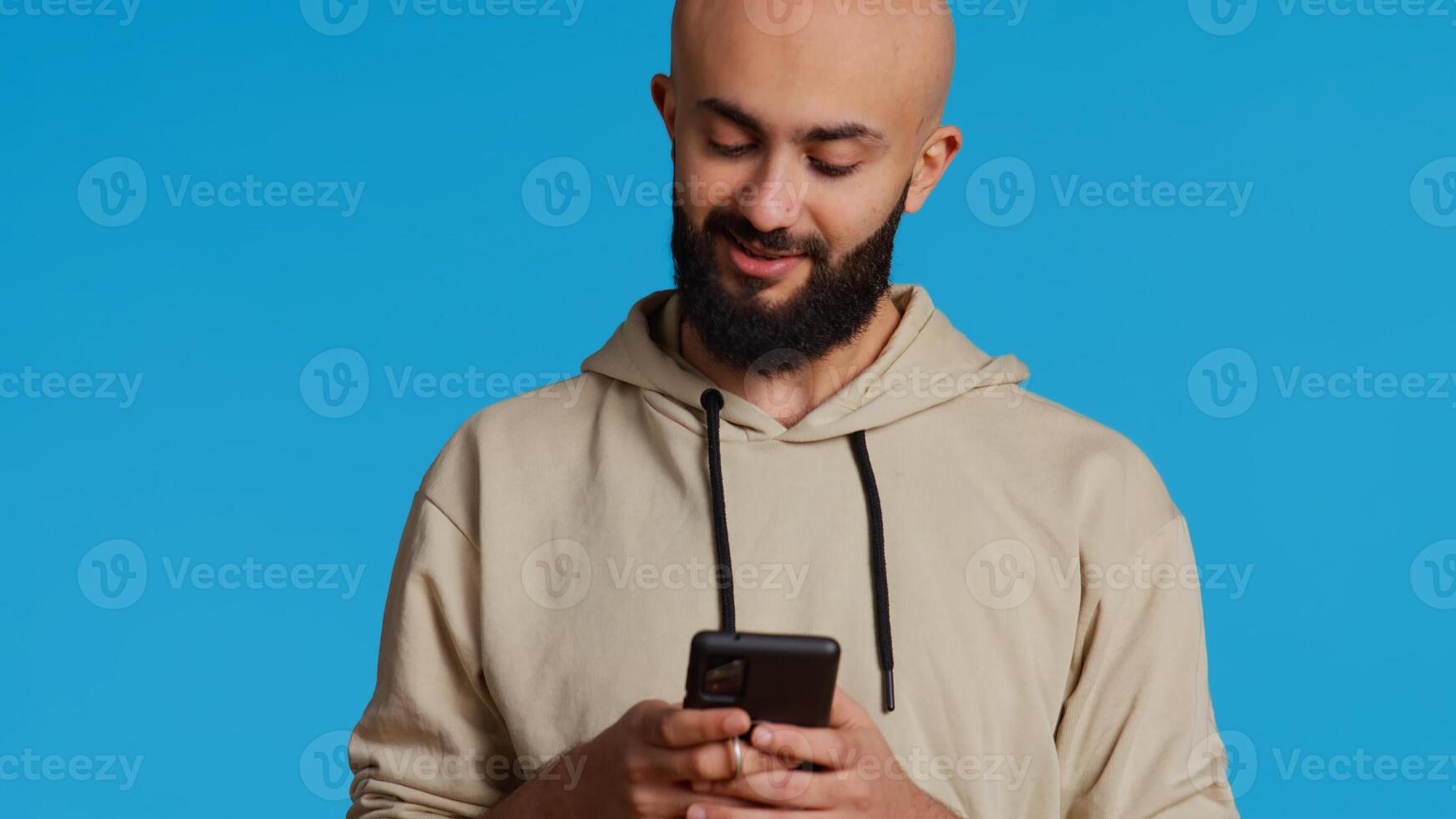 Arab person typing text messages on smartphone app, using mobile phone internet to check social media. Middle eastern guy browsing online websites in studio, blue backdrop. Camera 2. photo
