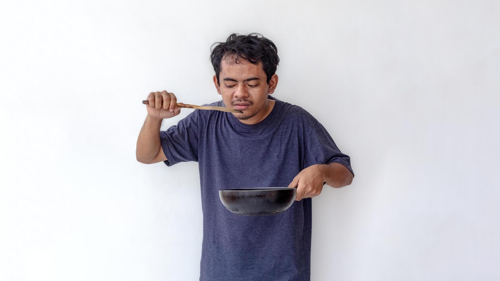 Adult man tasting food with a wooden spatula while holding a frying pan, isolated on a white background. Ideal for themes related to cooking, food preparation, and culinary skills. photo