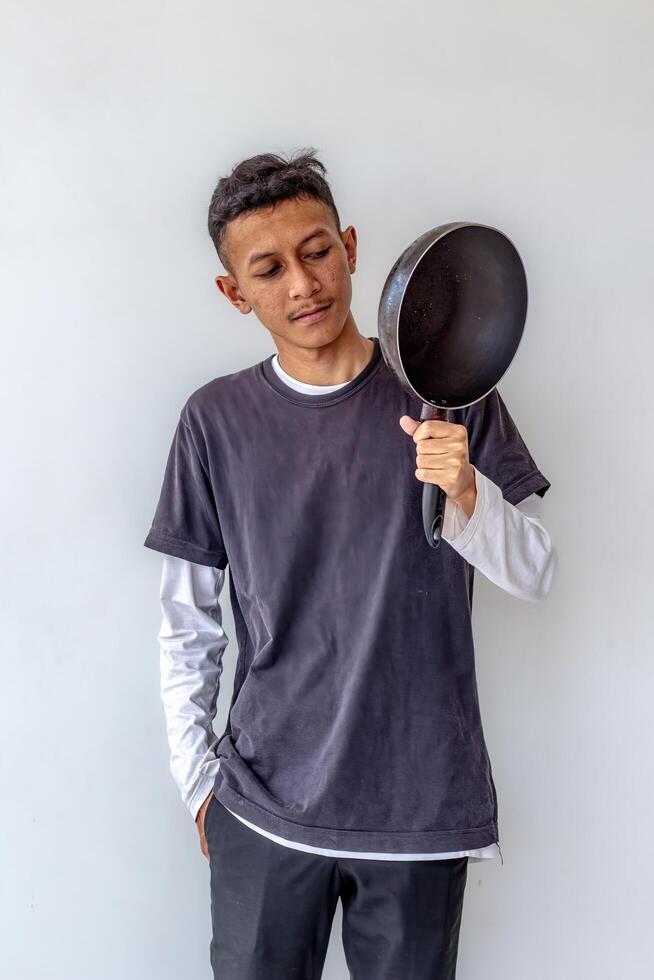 Young man holding and inspecting a frying pan, isolated on a white background. The image shows a man in a cooking stance, ideal for themes related to culinary skills, cooking, and kitchen activities. photo