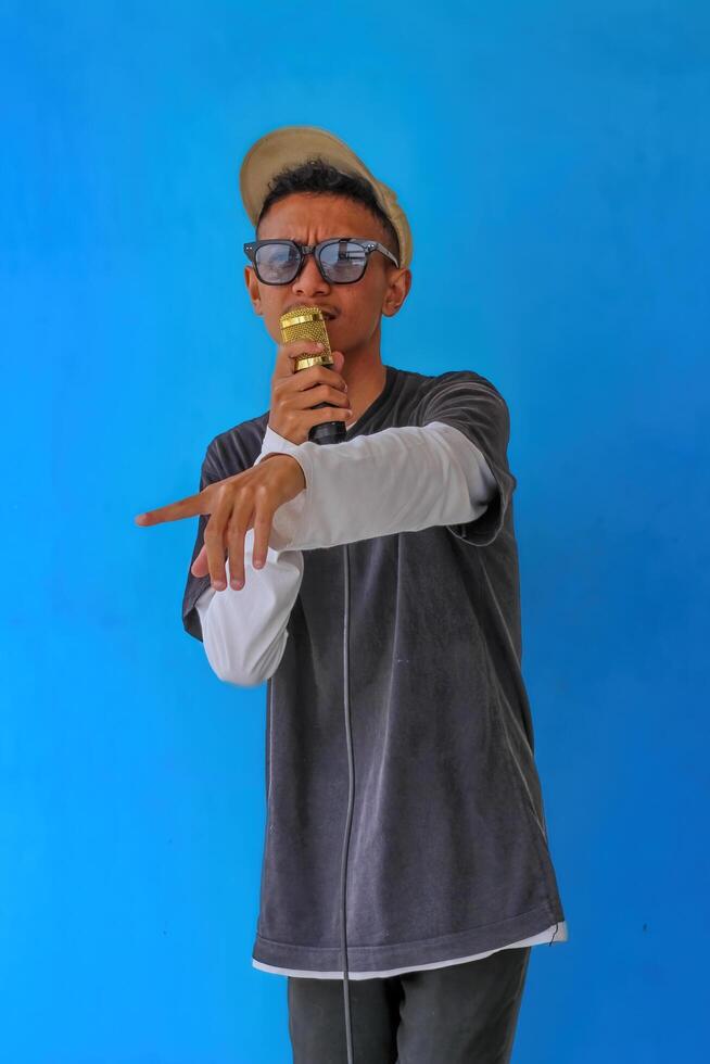 Young expressive singer happy man wearing gray t-shirt with glasses and hat singing song into microphone spreading hands isolated on plain light blue background studio. People's lifestyle concept photo