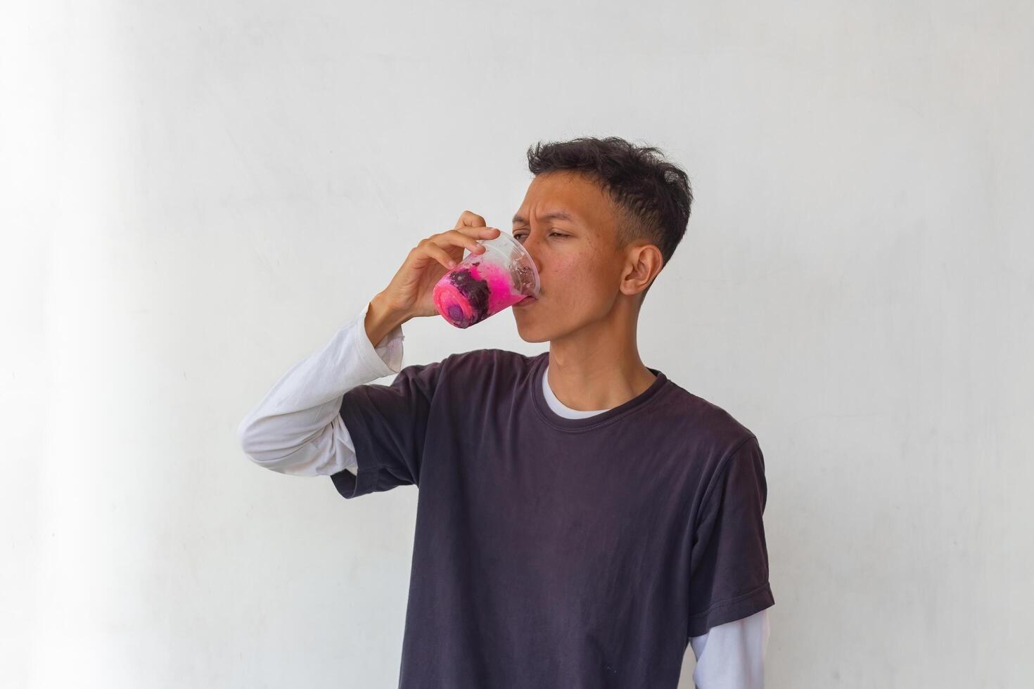 A young man wearing a gray t-shirt and glasses is enjoying an Indonesian mixed iced drink or holding a plastic cup isolated on white background. for food content and health, and diabetes concepts photo