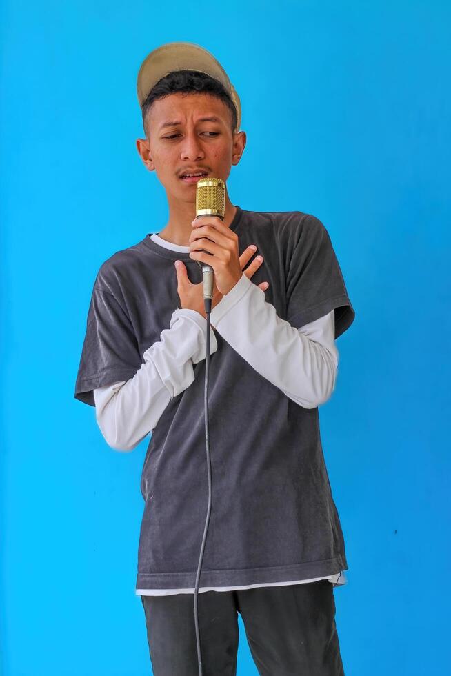 Young expressive singer happy man wearing gray t-shirt with glasses and hat singing song into microphone spreading hands isolated on plain light blue background studio. People's lifestyle concept photo