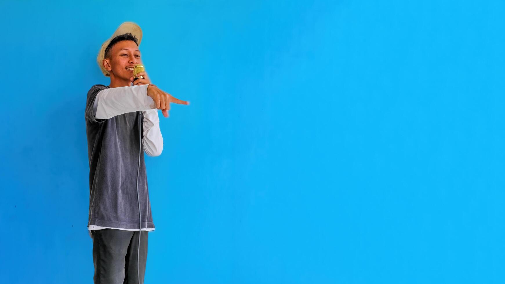 Young expressive singer happy man wearing gray t-shirt with glasses and hat singing song into microphone spreading hands isolated on plain light blue background studio. People's lifestyle concept photo