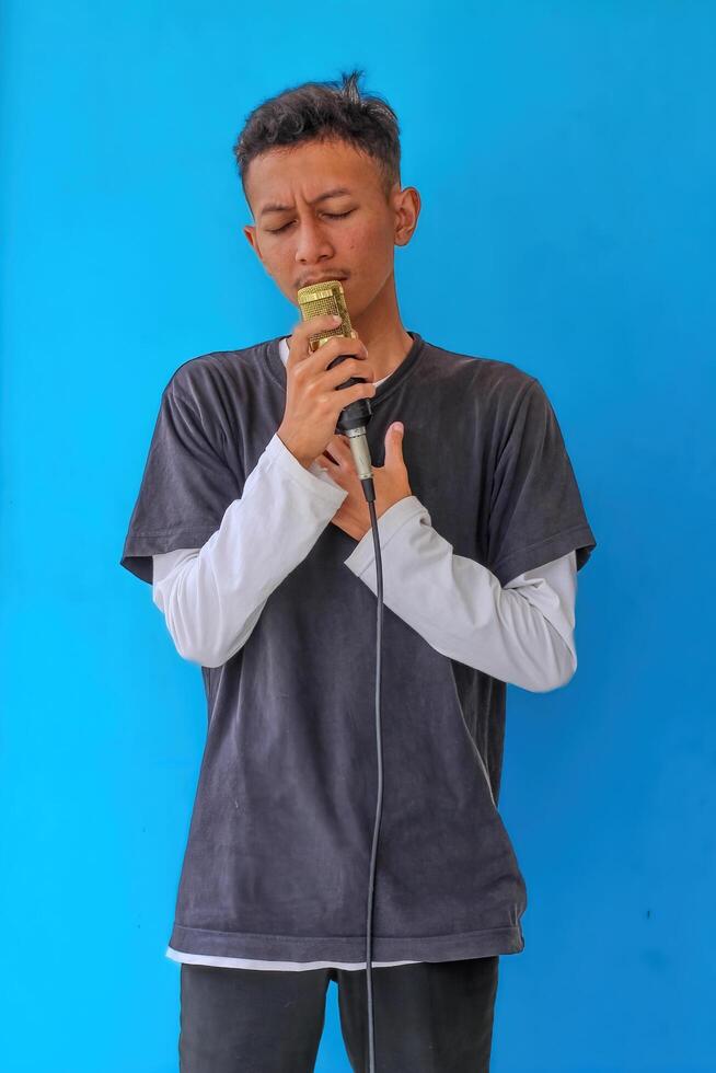Young expressive singer happy man wearing gray t-shirt with glasses and hat singing song into microphone spreading hands isolated on plain light blue background studio. People's lifestyle concept photo