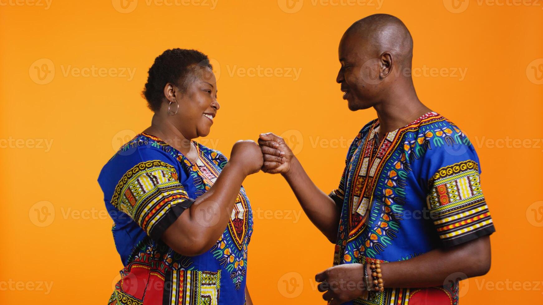 Young african american people greeting with fist bump on camera, saying hi to each other with modern gesture. Smiling husband and wife in traditional apparel posing in studio, cool salute. photo