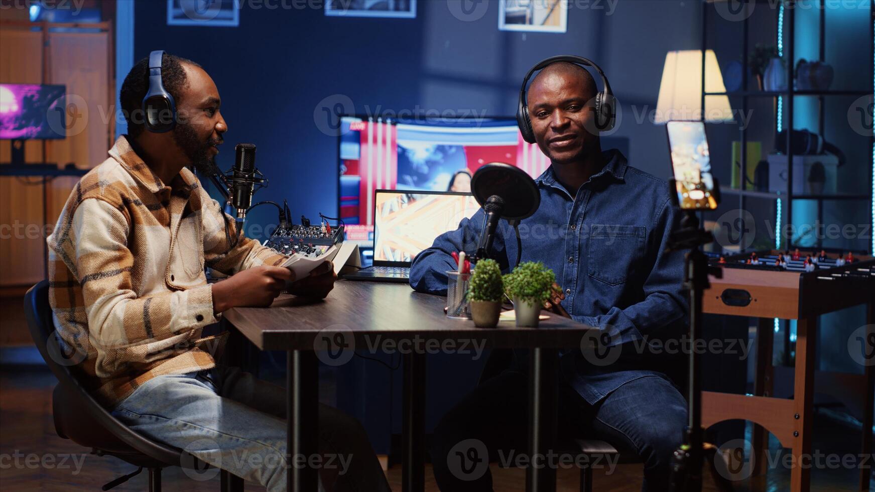 BIPOC host recording entertaining discussion with comedian during podcast filmed in apartment studio with phone on tripod stabilizer. Presenter uses cellphone camera to produce comedy internet show photo