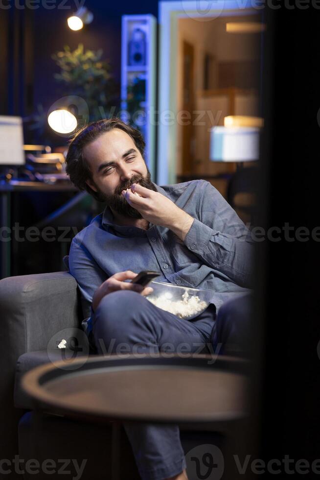 Overjoyed man sitting on couch laughing while watching funny comedy movie, eating popcorn. Upbeat person in apartment consuming snacks and being amused by hilarious joke in film scene photo
