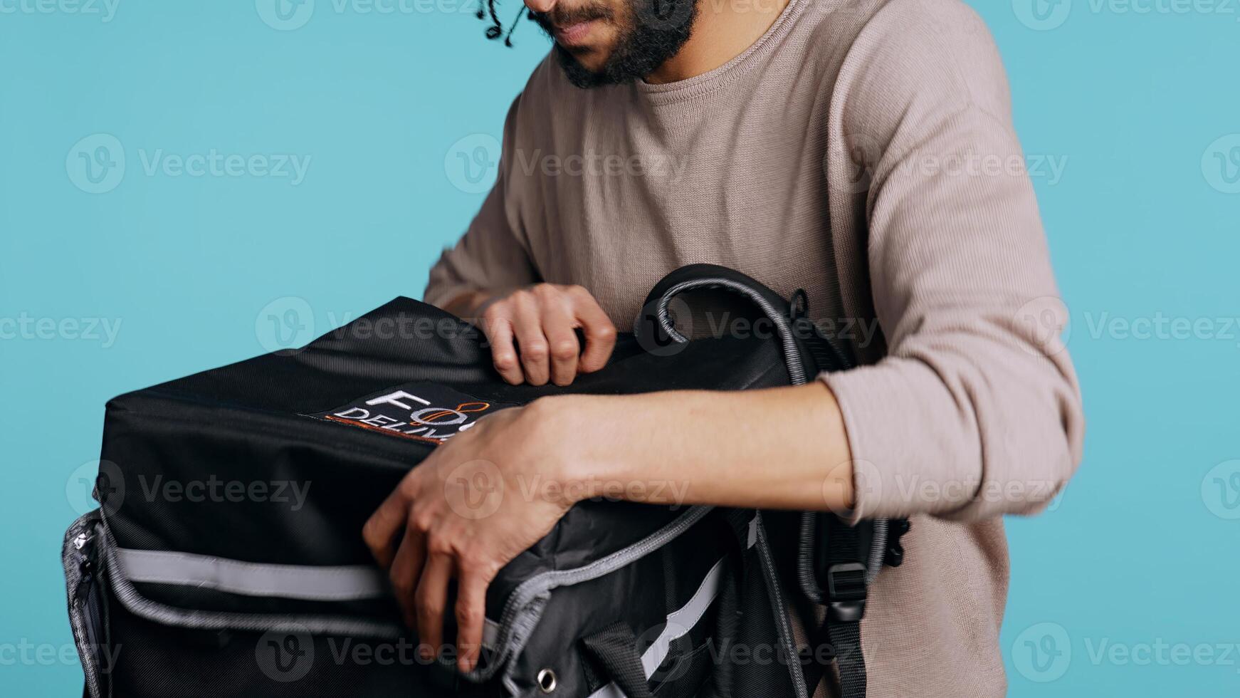 Portrait of pizza delivery guy taking takeaway paper lunchbag out of thermal bag. Middle Eastern courier getting meal out of food backpack, isolated over blue studio background, camera B photo