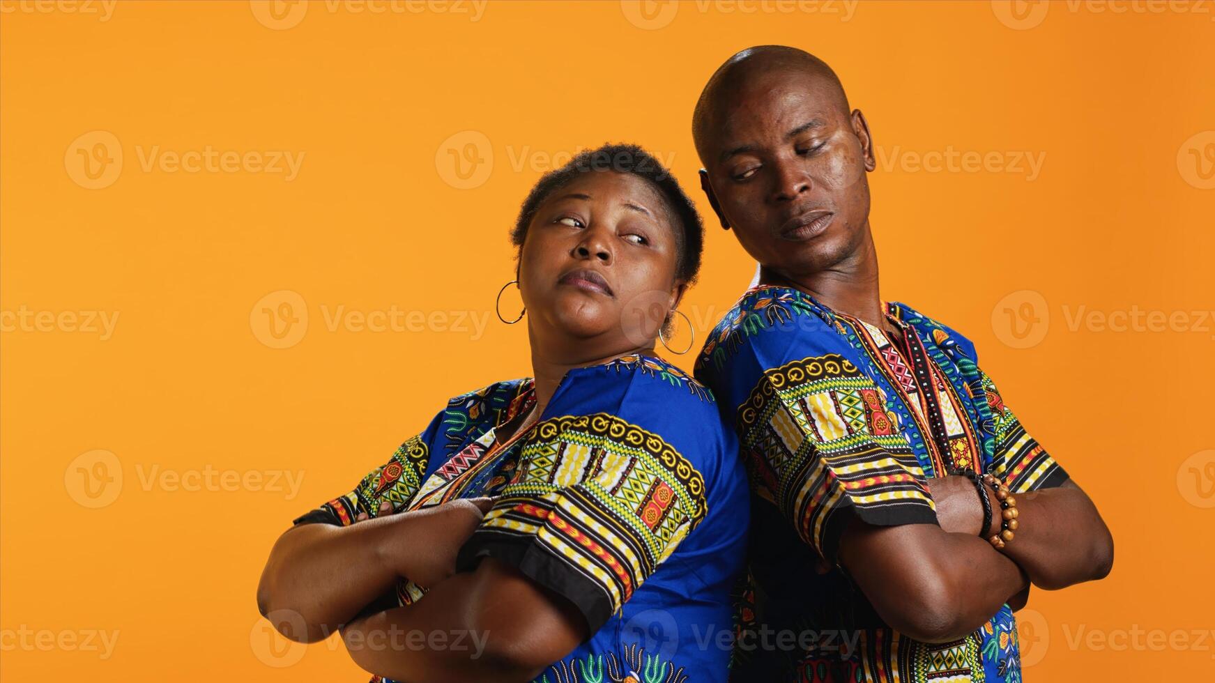 Carefree relaxed ethnic couple posing on camera, standing back to back and wearing traditional colorful clothes. African american people feeling confident and joyful together over orange background. photo