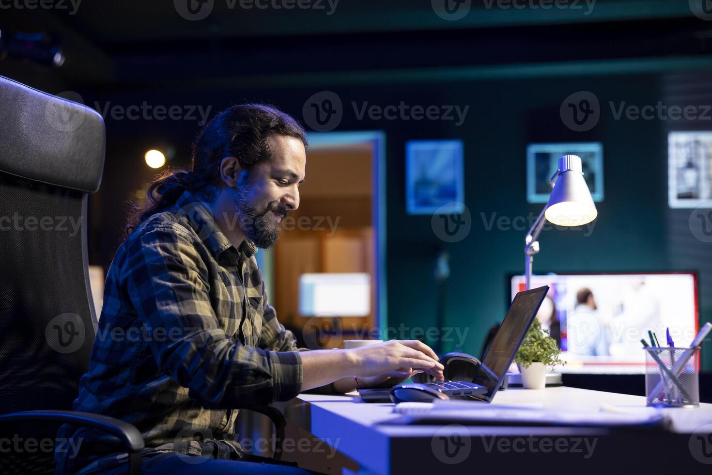 Smiling man at his living room desk, typing and browsing the internet on a laptop. Positive guy is concentrating on finishing several tasks as he sends emails on his electronic device. photo