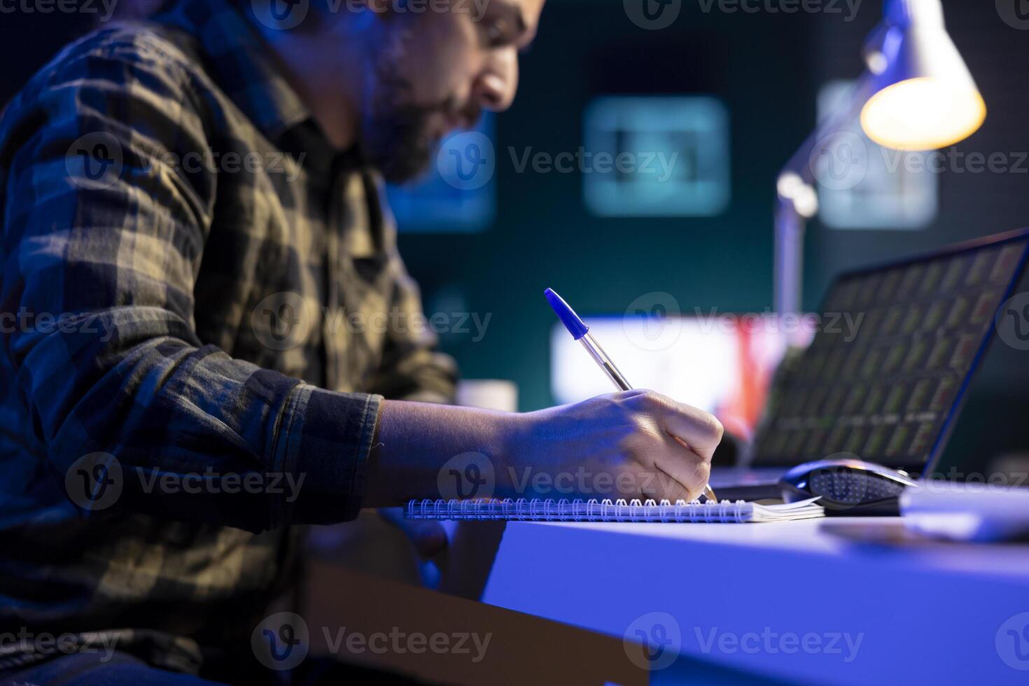 Diligent man multitasking, writing notes while reviewing forex chart on his laptop. Male freelancer working from home, using his notebook and checking status of stock market trend values on device. photo