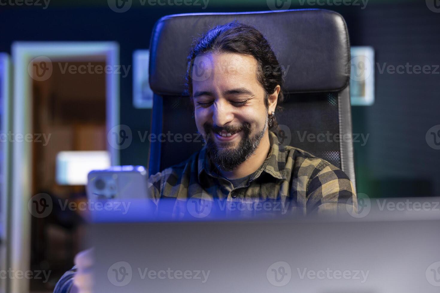 Male freelancer taking a break, cheerfully using his smartphone to watch funny online content. Positive young man sitting at desk laughing while texting on his mobile device, surfing the net. photo