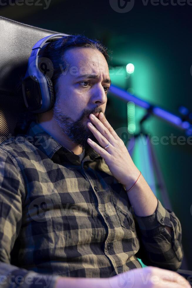 Shocked man sitting at a desk, watching a movie on portable computer while wearing headphones. Young guy listening to a podcast on wireless headset while surfing the net on laptop. photo