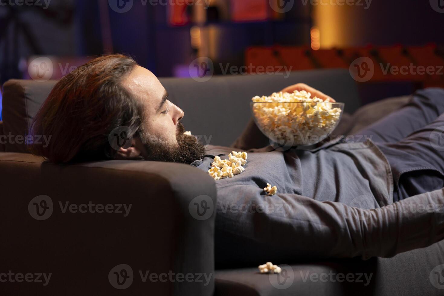 Man sleeping with bowl of popcorn on him after watching cable programs on TV at home. Person falling asleep on couch in living room after looking at television series late at night photo