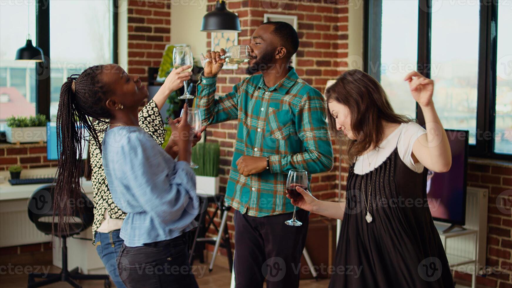 Energetic group of friends dancing at apartment party, having fun together while drinking wine. Multiethnic people doing dance moves on lively music in brick wall living room photo