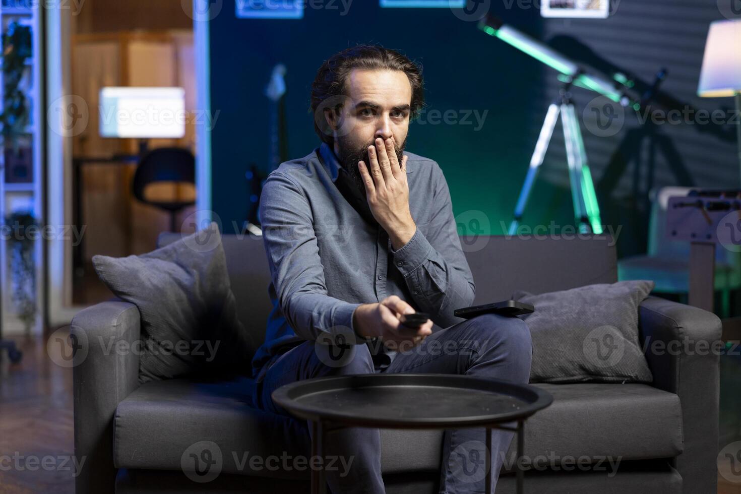 Man gasping while watching horror movie on streaming service, enjoying time off from work, holding remote control. Person covering mouth in disbelief, surprised by film scene broadcasted on TV photo