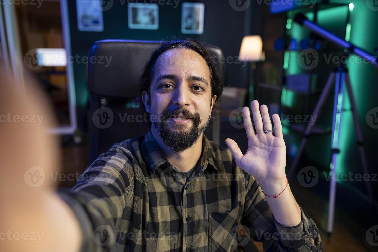 Male influencer waving his hand towards camera, making a daily vlog at home. Smiling young man having call with friends using modern technology. photo