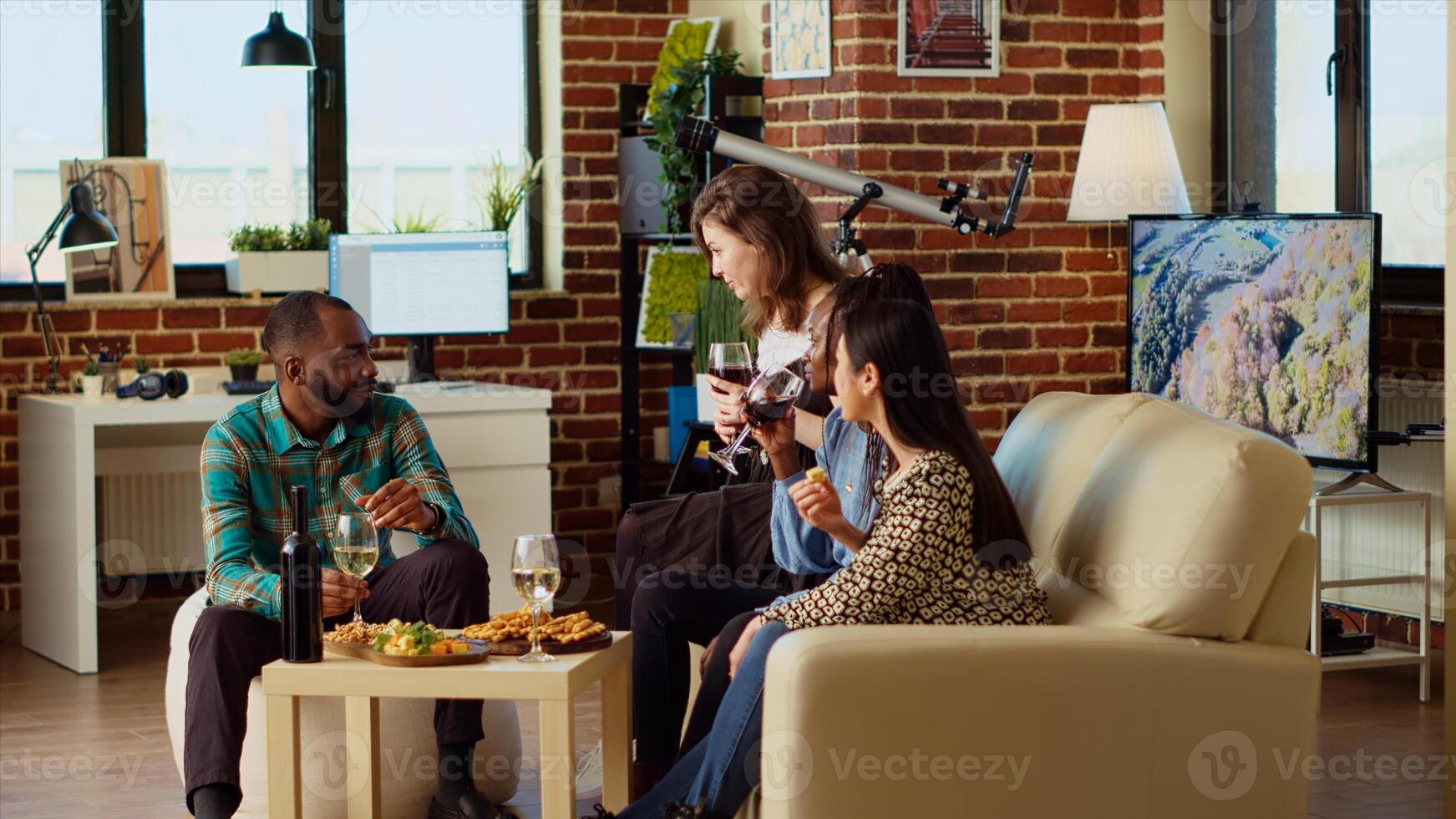 Multiracial group of people having entertaining conversation during their friendship celebration at home. Joyful friends having reunion gathering party in cozy living room, enjoying alcoholic drinks photo