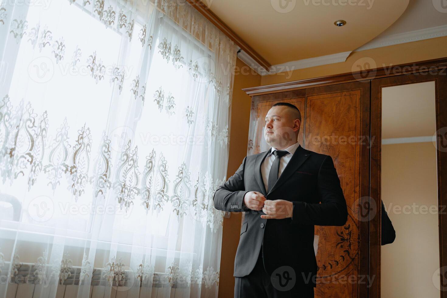 A man in a suit is standing in front of a window, adjusting his tie. Concept of formality and professionalism, as the man is dressed in a suit and tie photo