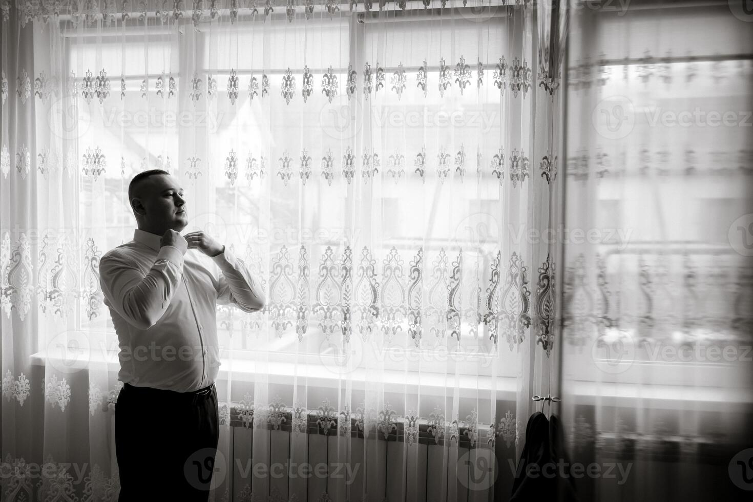 A man is getting dressed in front of a window. The curtains are white and the window is letting in some light. The man is wearing a white shirt and black pants photo