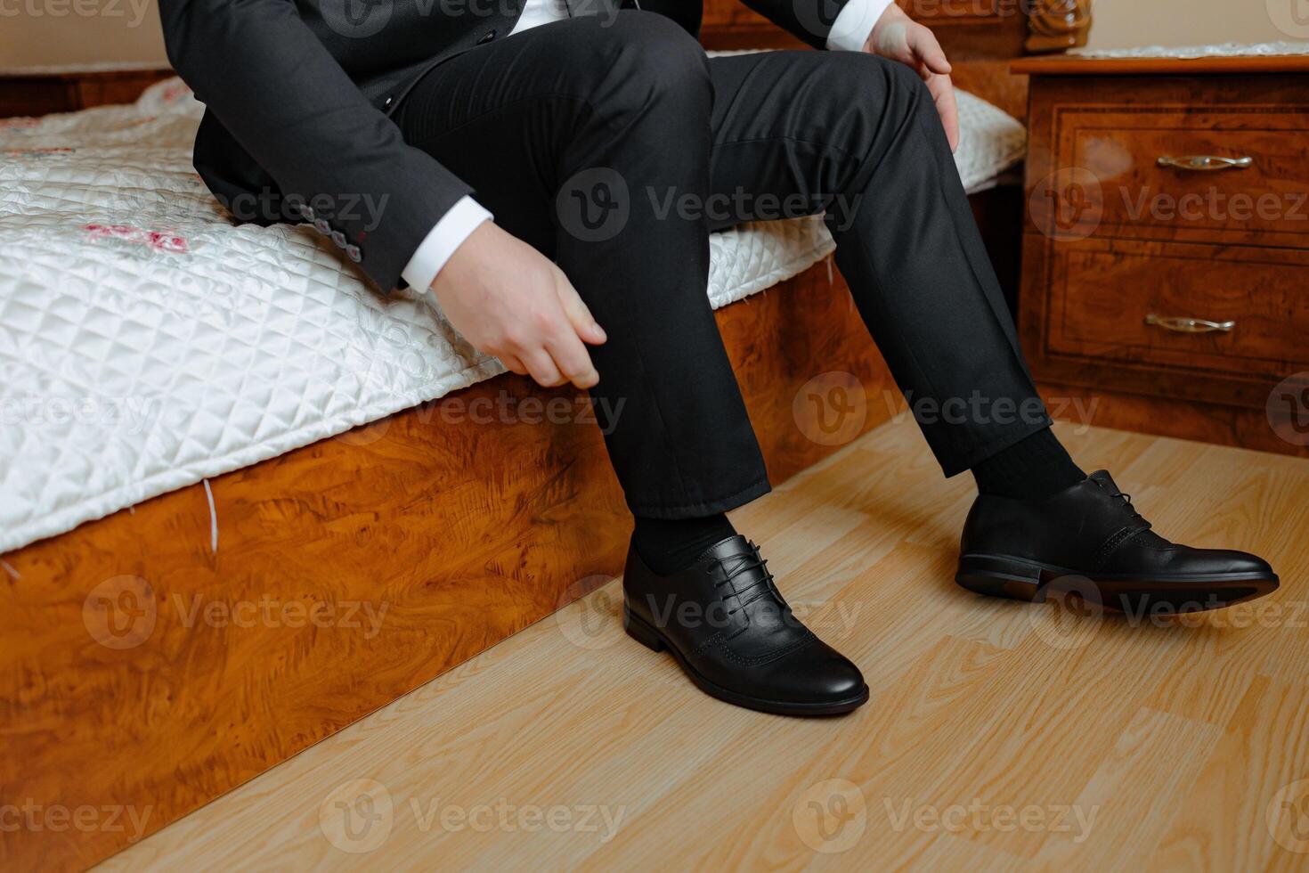 A man in a suit is sitting on a bed with his feet up. He is wearing black dress shoes and is adjusting them. Concept of relaxation and comfort, as the man is taking a break from his formal attire photo