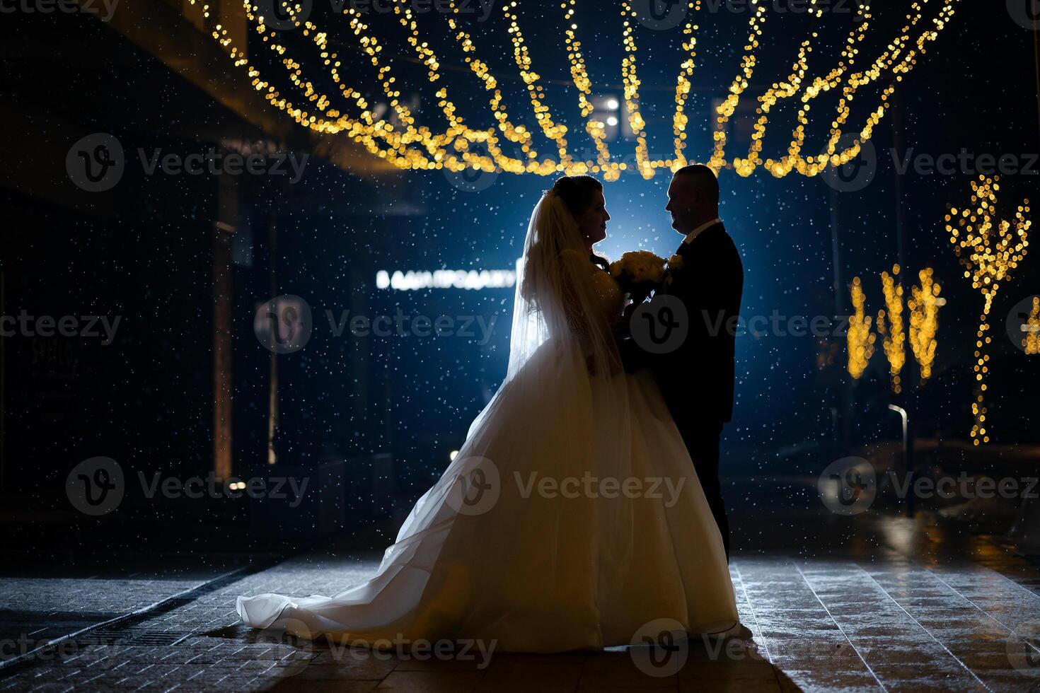 A bride and groom are standing in the rain, with the bride holding a bouquet. The scene is dark and moody, with the couple looking into each other's eyes photo
