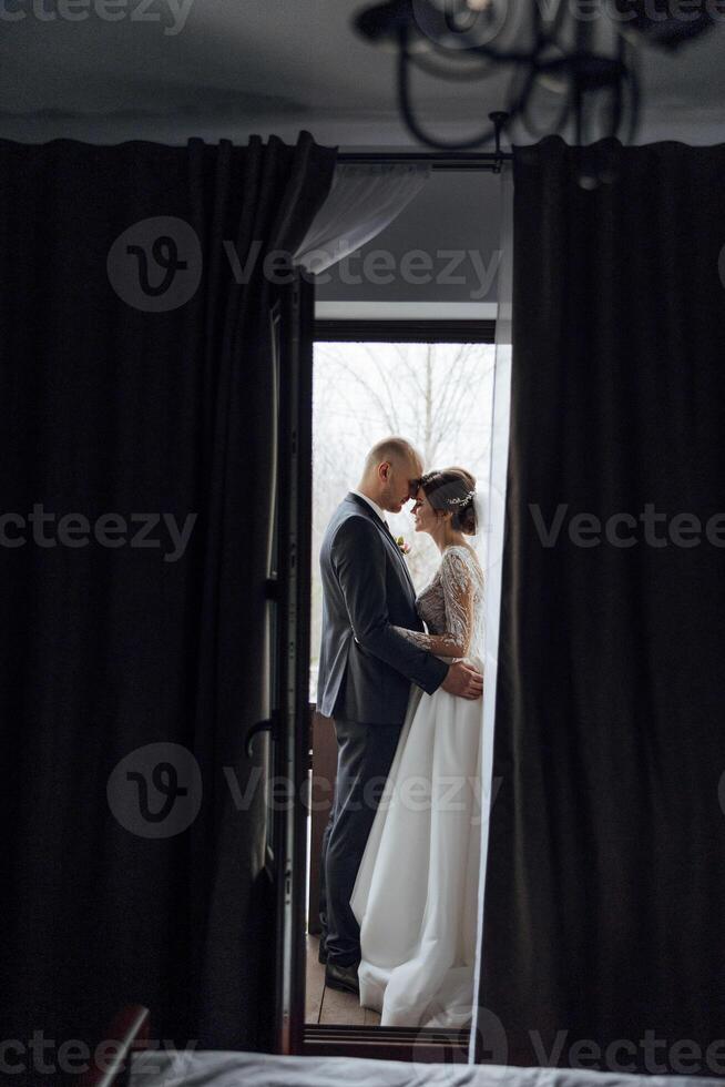 A bride and groom are standing in front of a window, with the bride wearing a white dress photo