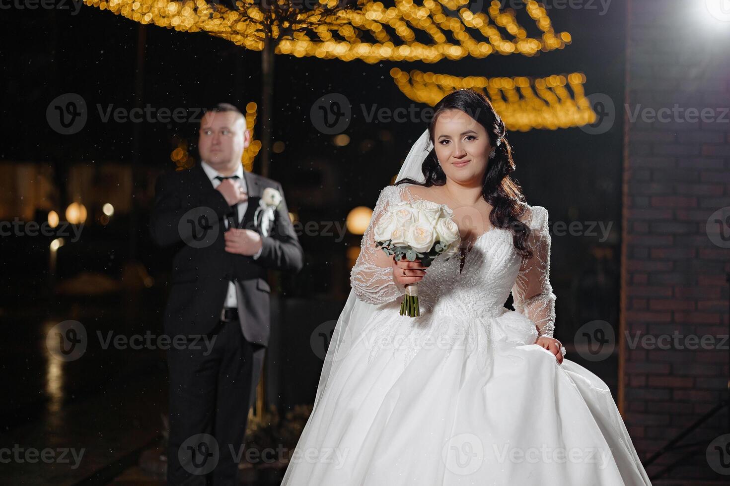 A bride and groom are posing for a picture in front of a brick building. The bride is holding a bouquet of flowers and the groom is standing behind her. Scene is romantic and happy photo
