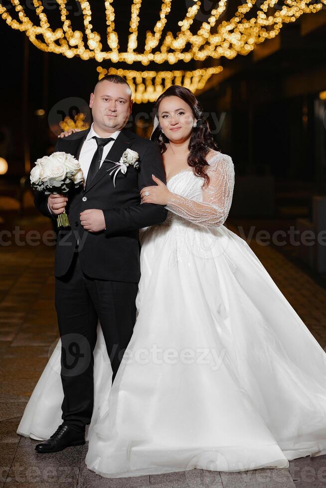 A bride and groom are posing for a picture in front of a lighted sign. The bride is wearing a white dress and the groom is wearing a suit. The couple is holding a bouquet and a tie, respectively photo