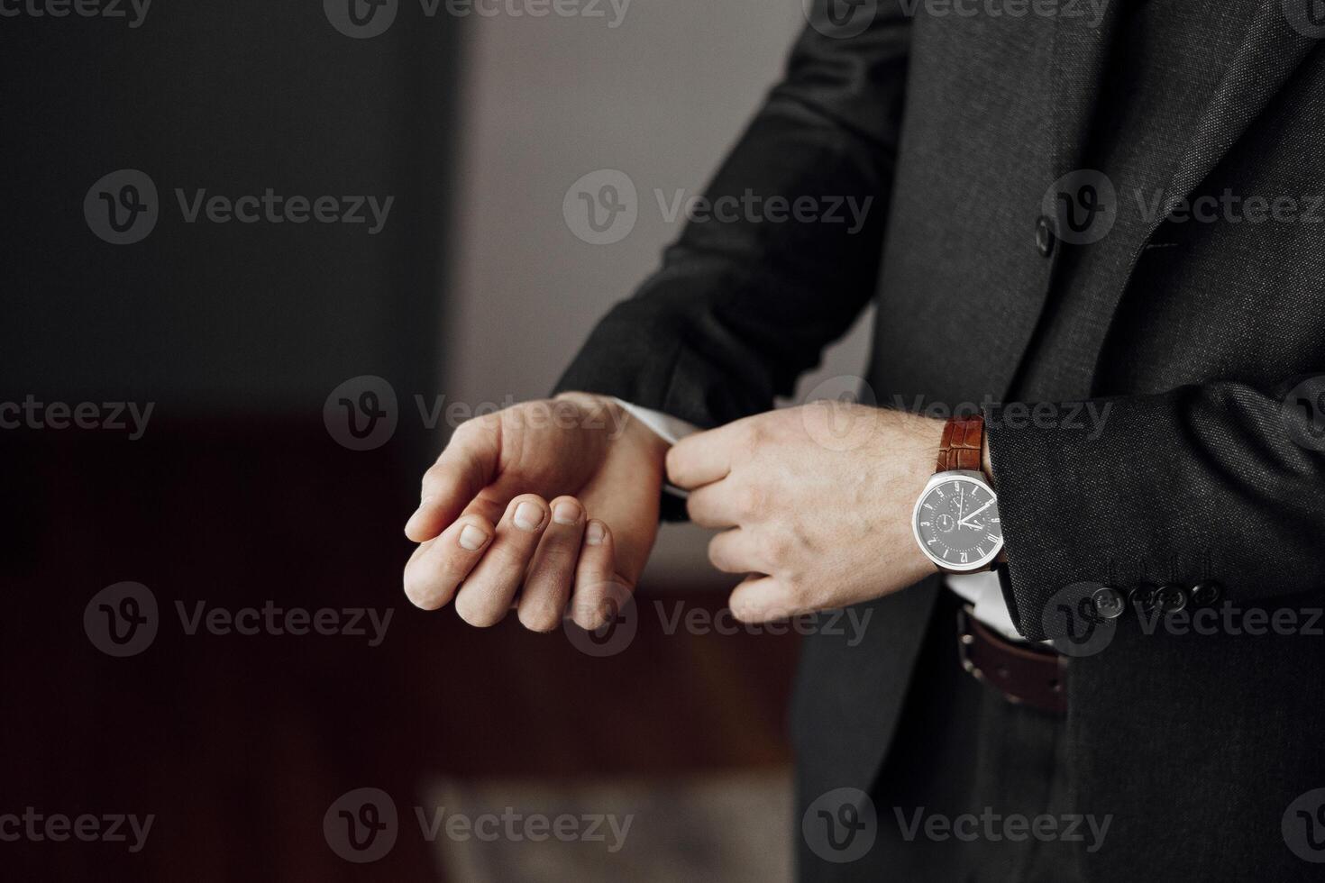 A man in a suit is adjusting his watch. The man is wearing a black suit and a brown watch band photo