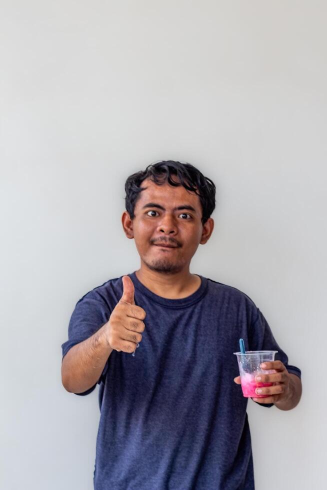 A young adult wearing a gray t-shirt and glasses is enjoying an Indonesian mixed iced drink or holding a plastic cup isolated on white background. for food content and health, and diabetes concepts photo