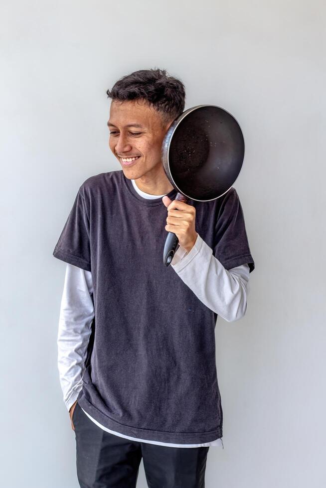 Young man lifting a frying pan and smiling, isolated on a white background. The image shows a cheerful cook in action, perfect for themes related to cooking, kitchen joy, and culinary enthusiasm. photo
