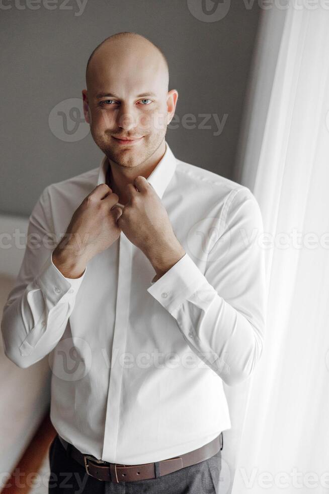 A man in a white shirt is standing in front of a window, adjusting his collar. The image has a casual and relaxed mood, as the man is getting ready for a day at home or work photo
