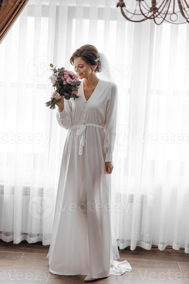A bride is holding a bouquet of flowers and standing in front of a window. Scene is romantic and elegant, as the bride is dressed in a long white dress and is surrounded by the beauty of the flowers photo