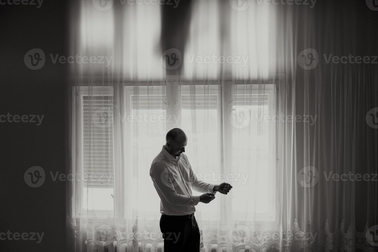 A man in a white shirt is standing in front of a window, adjusting his tie. The image has a moody and somewhat somber feel, as the man is in a reflective or contemplative state photo
